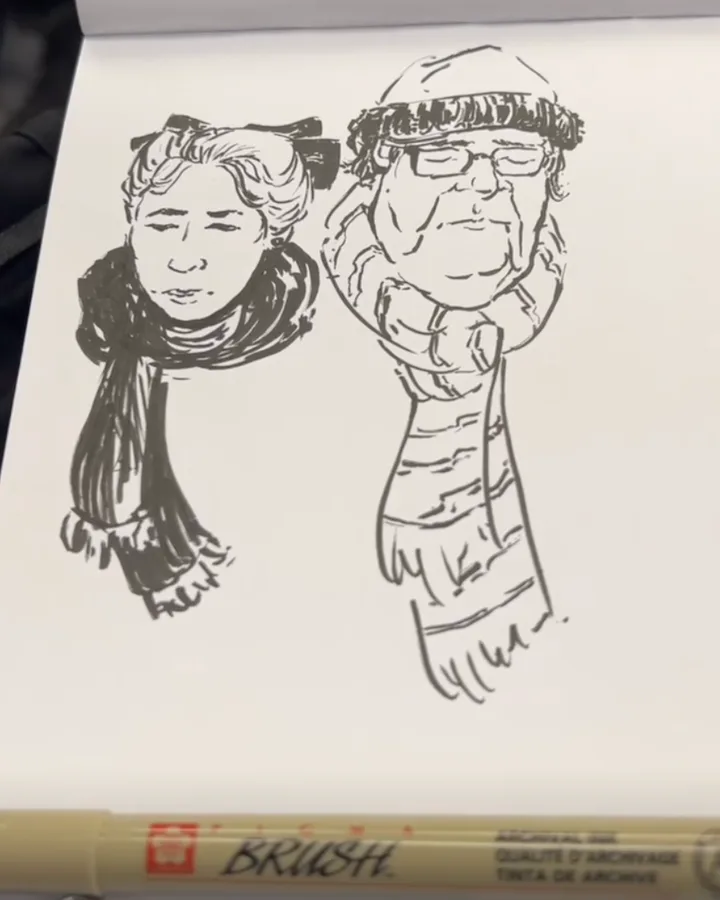 Brush-pen sketch on a white pad of two seated passengers: a woman with grey hair pulled up under a bow and a dark scarf, and a man in a knitted beanie, glasses, and a striped scarf. The pen lies across the bottom of the page.