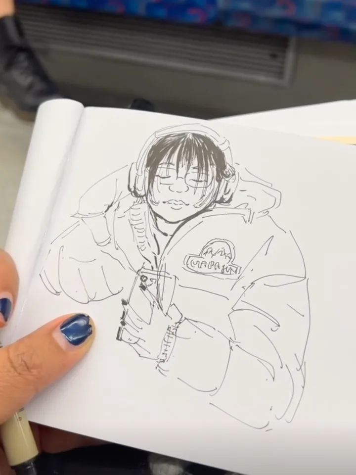 Fineliner sketch of a passenger with a dark bob, eyes closed under over-ear headphones, wrapped in a puffer jacket and holding a phone. A hand with blue nails holds the sketchbook open; the patterned seat fabric shows above.