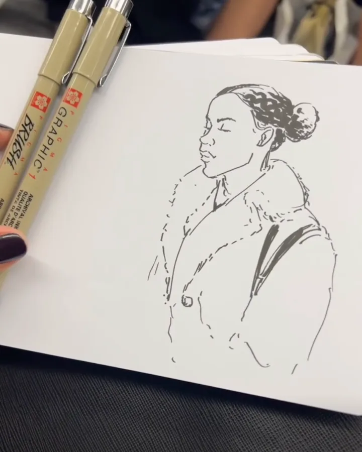 Ink profile of a woman with her hair braided back into a high bun, wearing a collared coat with a shoulder bag strap. Two fineliners are clipped along the left edge of the sketchbook.